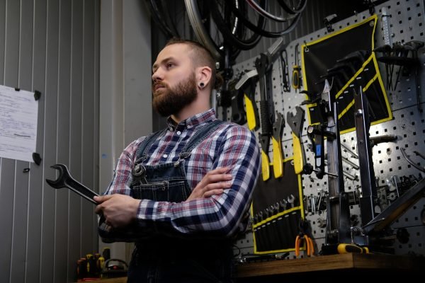 Portrait of a handsome stylish male with beard and haircut wearing a flannel shirt and jeans coverall, holds steel wrench, standing in a workshop against wall tools.